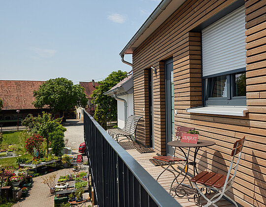 Ferienwohnung Balkon Lemp’s Genussgarten Balkon mit Holzwand, zwei Stühlen und Tisch, Blick auf Garten mit Pflanzen und Gebäuden im Hintergrund an sonnigem Tag.