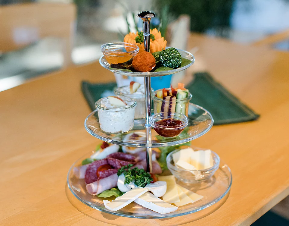 Lemp's Genussgarten Gärtnerfrühstück Dreistöckiges Glas-Etagere mit verschiedenen Käsesorten, Wurst, Frischkäse, Dips und dekorativen herzhaften Snacks auf einem Holztisch.