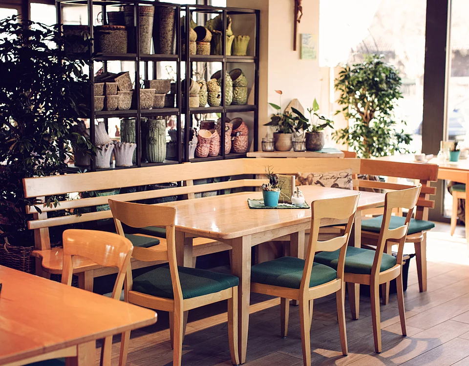Lemp’s Genussgarten Café Innenansicht eines Cafés mit Holztischen, Stühlen mit grünen Sitzpolstern und Regalen mit Körben und Pflanzen im Hintergrund.