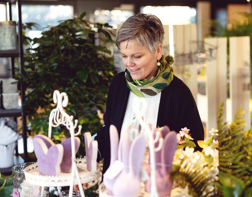 Lemp’s Genussgarten Floristik Frau mit kurzem Haar schaut lächelnd auf bunte Osterdekoration in einem Laden mit Pflanzen und frühlingshaften Motiven.