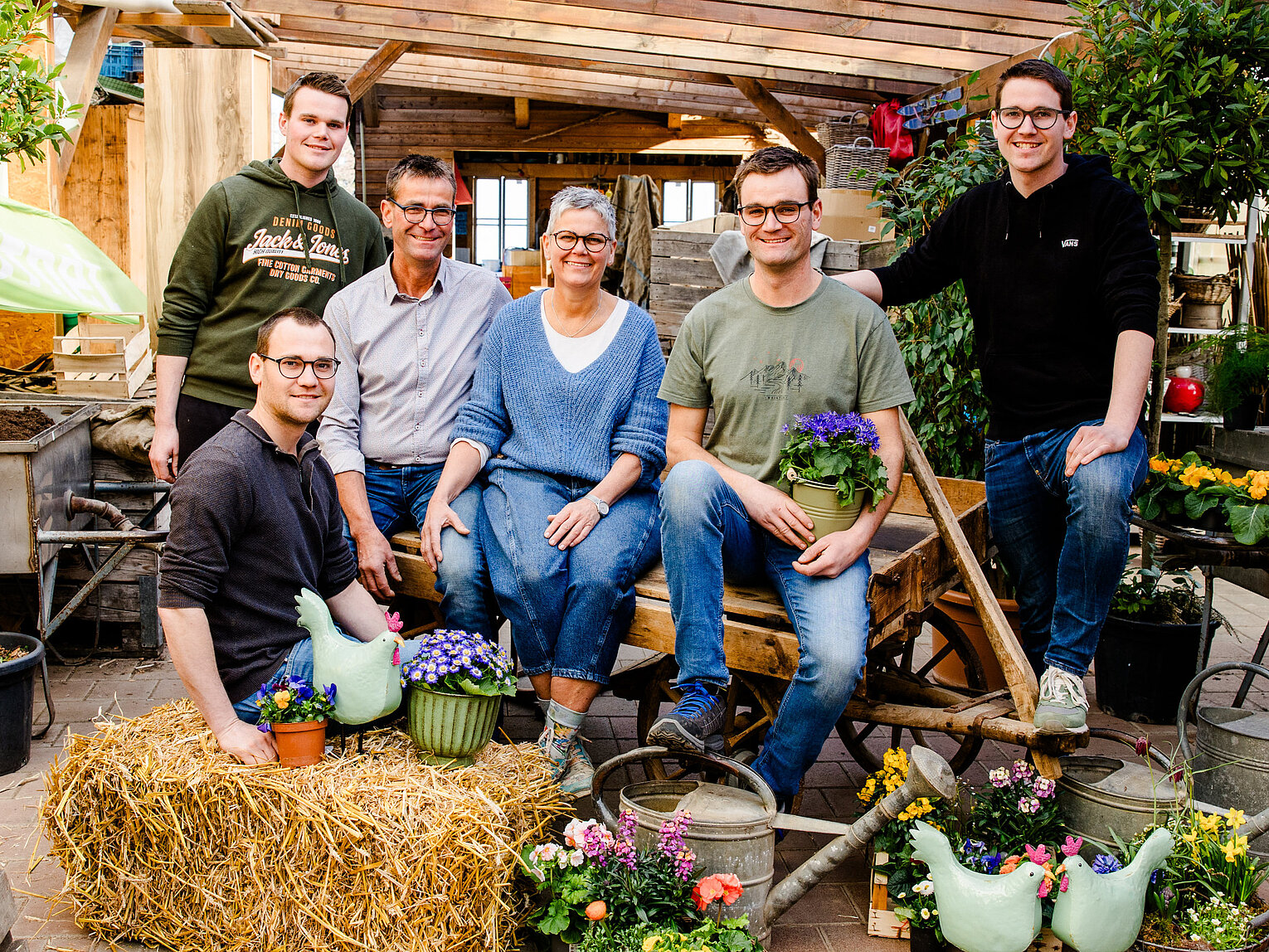 Team von Lemp’s Genussgarten Sechs Menschen, vier Männer und zwei Frauen, sitzen und stehen in einem Gewächshaus um eine Heuballen und bunte Blumentöpfe. Sie lächeln und wirken entspannt in einer Gartenumgebung mit Pflanzen und Gartengeräten.